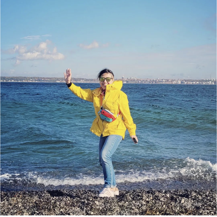 Yi Ming He waving on a beach, wearing a yellow jacket and sunglasses